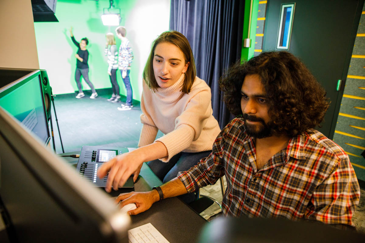 Students working at a computer with students performing in front of a green screen in the background.